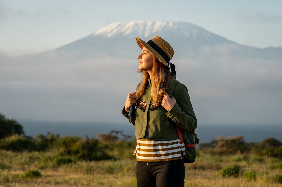 A beautiful young girl in a hat stands against the backdrop of the Kilimanjaro volcano and looks away