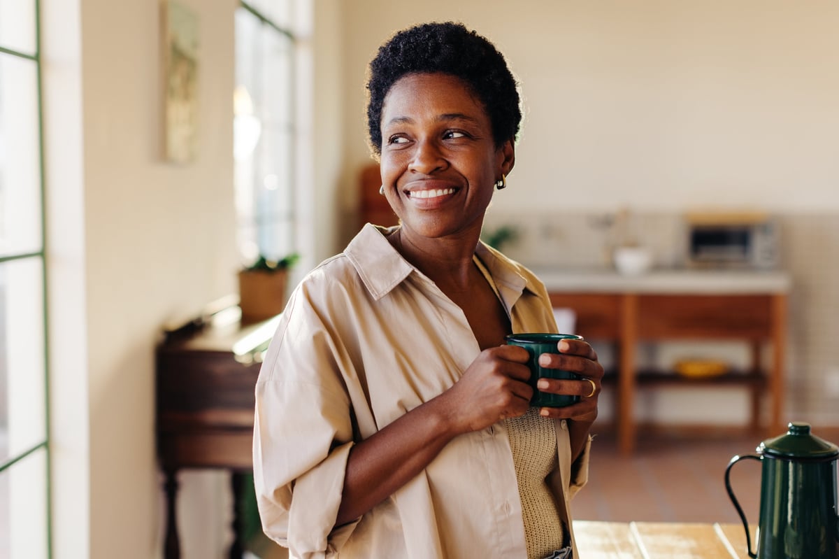 Happy Brazilian woman looking outside the window in her home kitchen, smiling and holding an enamel mug of plain coffee