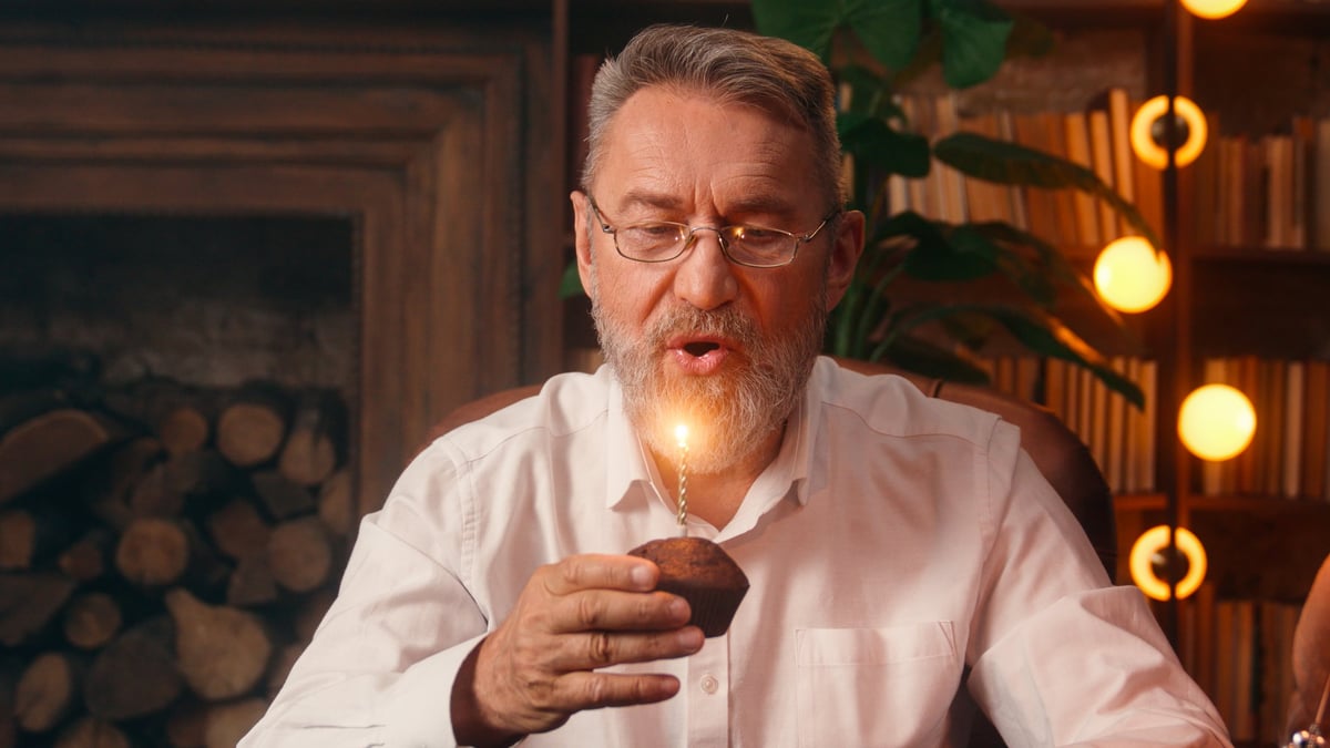 Happy joyful elderly man blowing out a candle on a birthday cake at home, celebrating with a smile