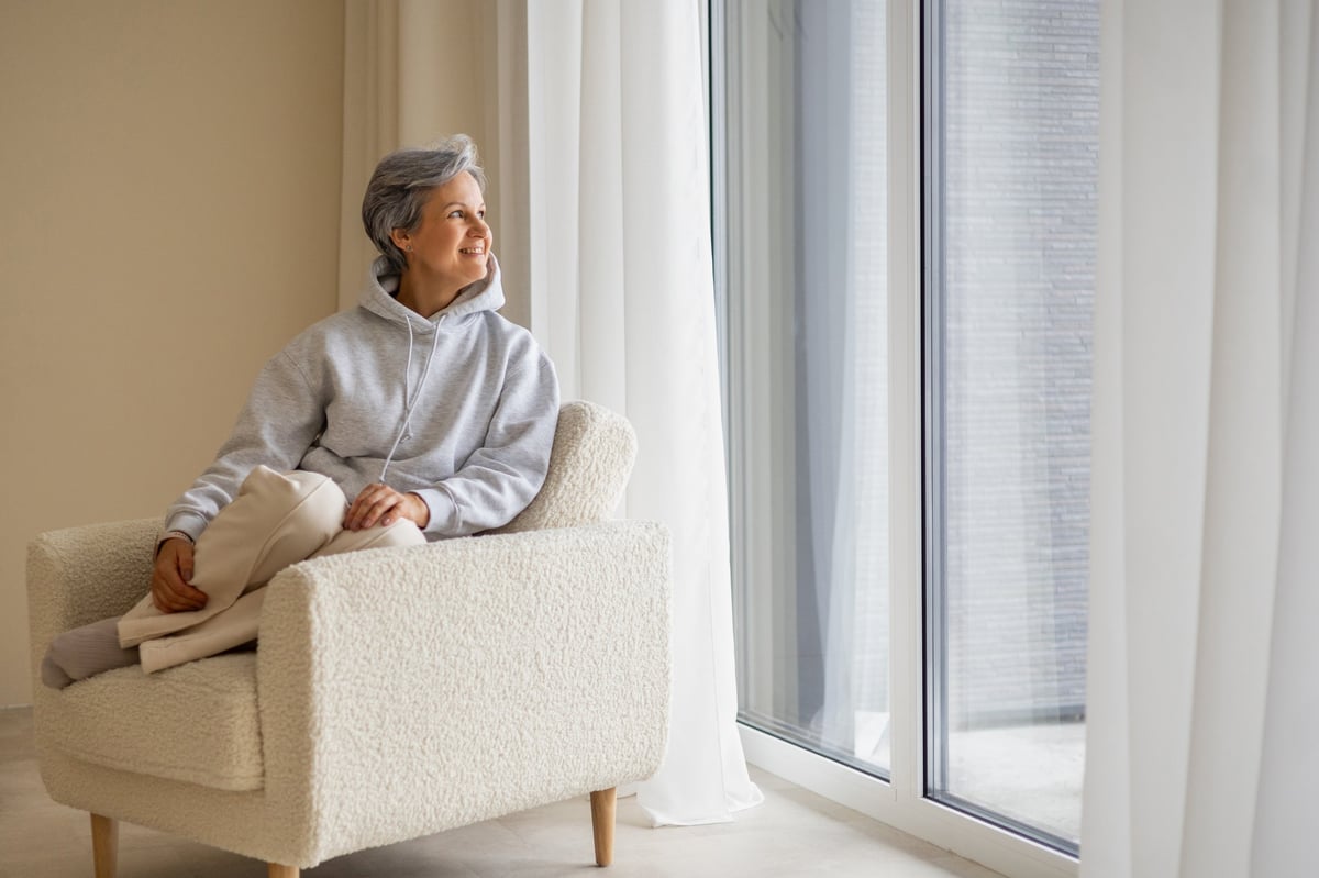 Calm senior woman with short gray hair in light hoodie sits barefoot in a soft beige chair and looks out the window. Cozy minimal interior, soft daylight, peaceful lifestyle.