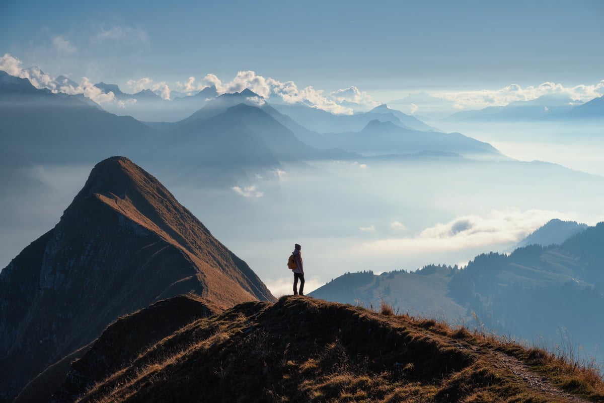 Woman with backpack standing on a mountain ridge above clouds in the Swiss Alps at sunset