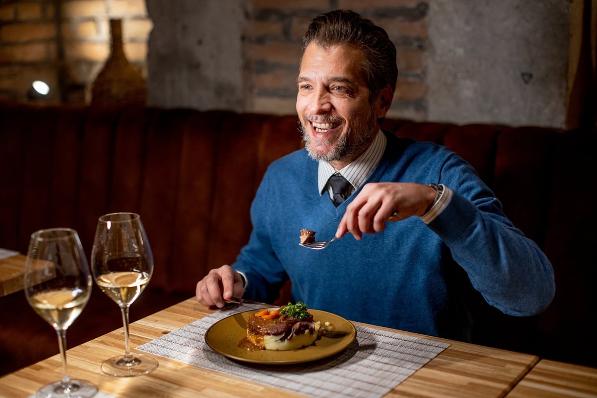 Man sits at a table in a restaurant. He smiles while holding a fork and looks at his meal. Two glasses of wine sit on the table beside him.