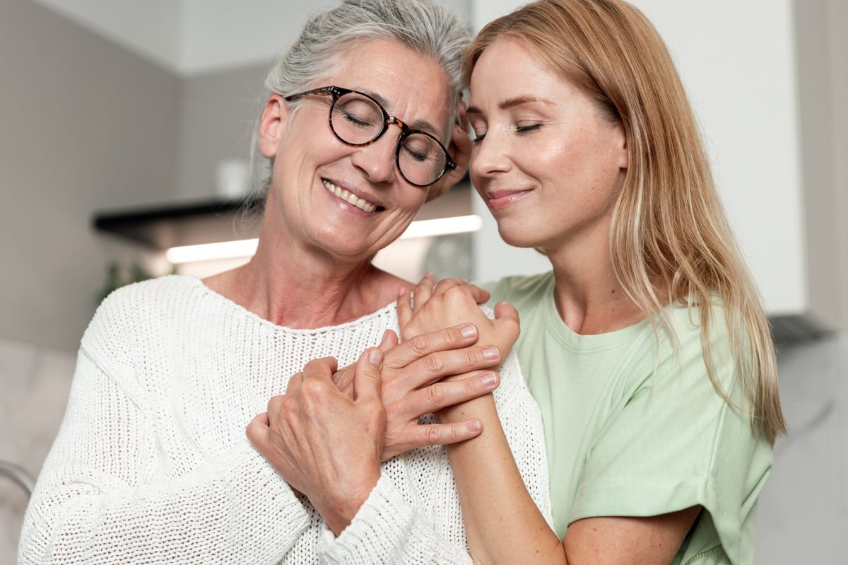 In a cozy apartment living room, an elderly mother and her adult daughter smile and hold hands