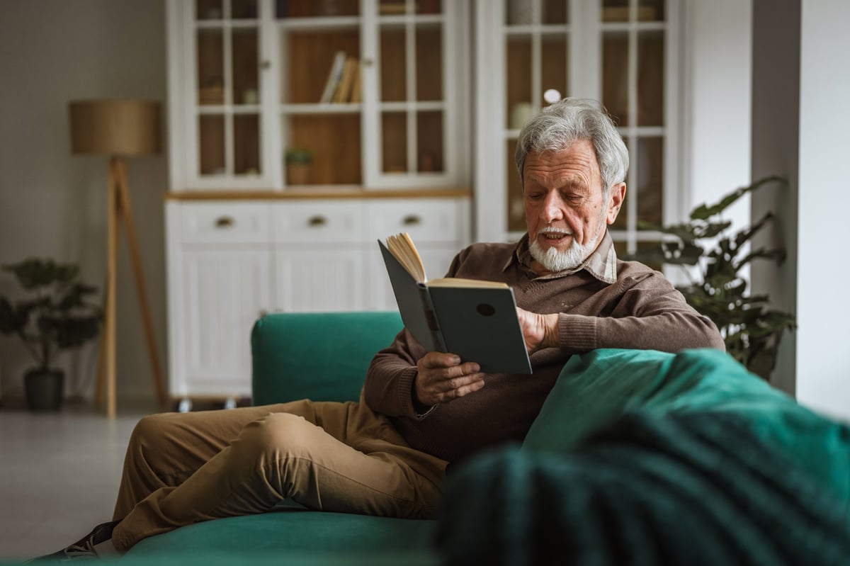 Senior man with gray hair and beard relaxing on a sofa at home, absorbed in a book and enjoying peaceful, thoughtful leisure in a comfortable living room