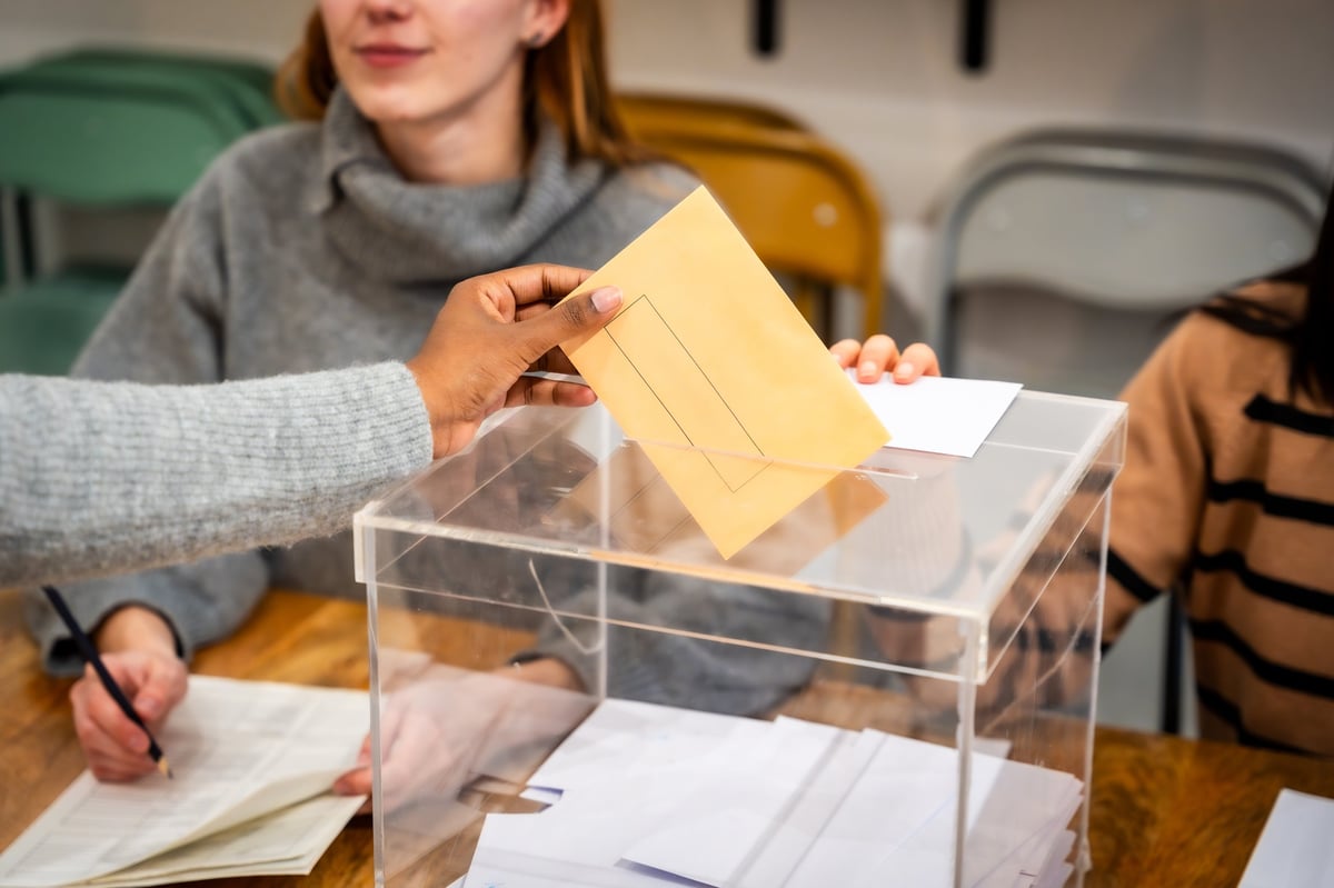 Voter's hand inserting a ballot envelope into a clear plexiglass ballot box during an election, representing democracy, civic duty, and the electoral process