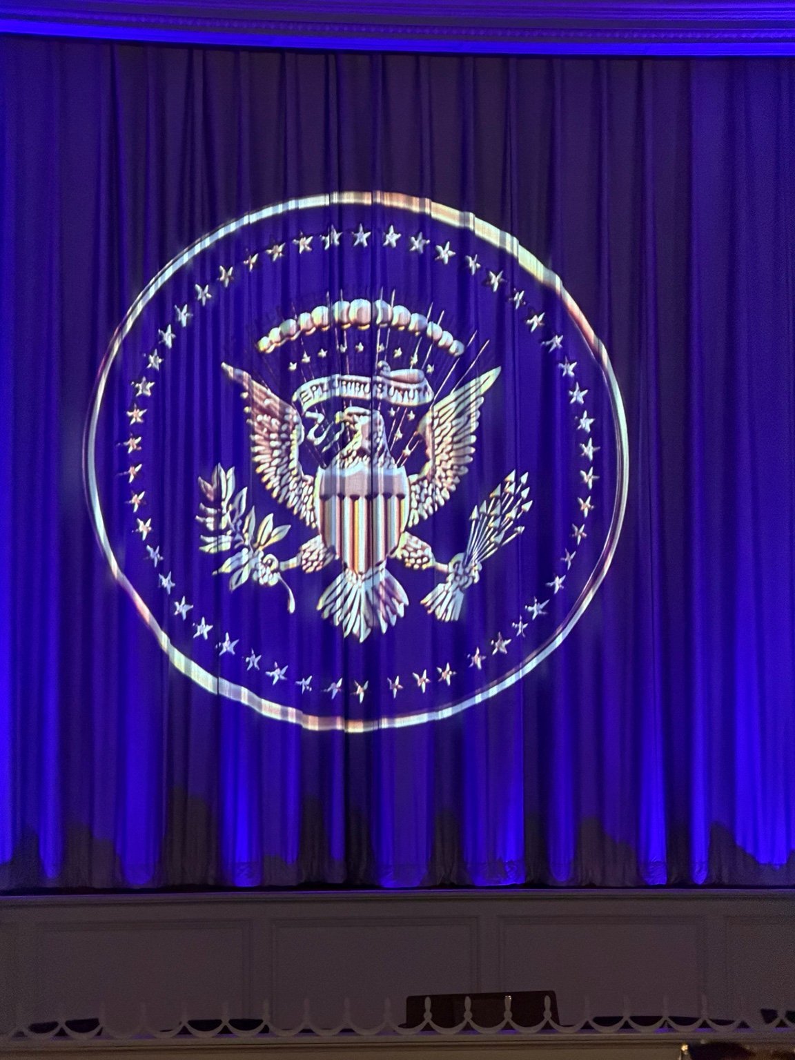 Presidential Seal illuminated in white neon against deep blue curtains with blue stage lighting