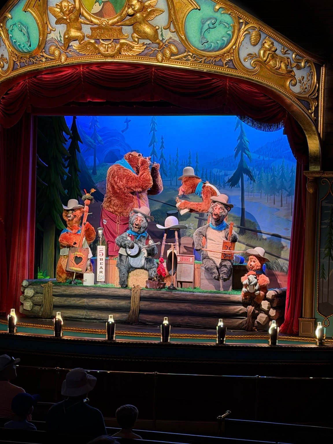 Ornate theater stage with red velvet curtains displaying a puppet show scene featuring cowboys and forest backdrop