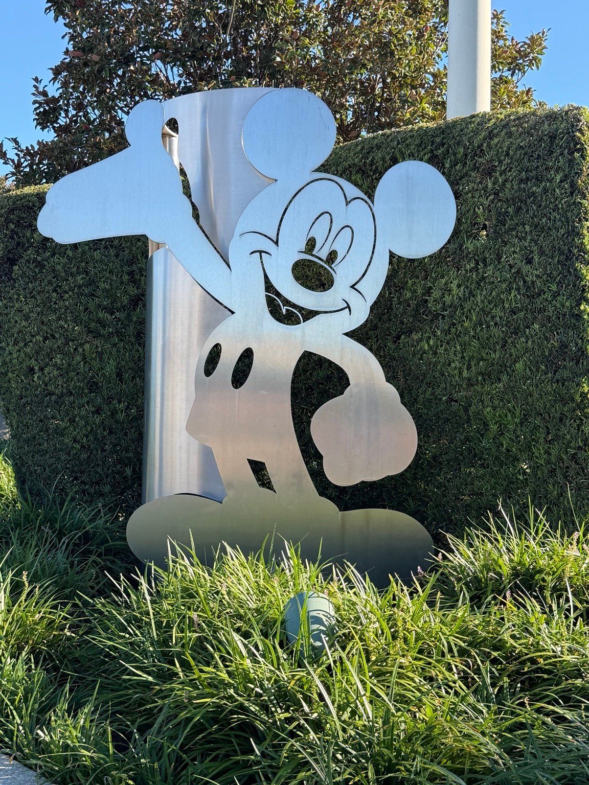 Large white Mickey Mouse sculpture with black gloves and shorts against green hedges and blue sky